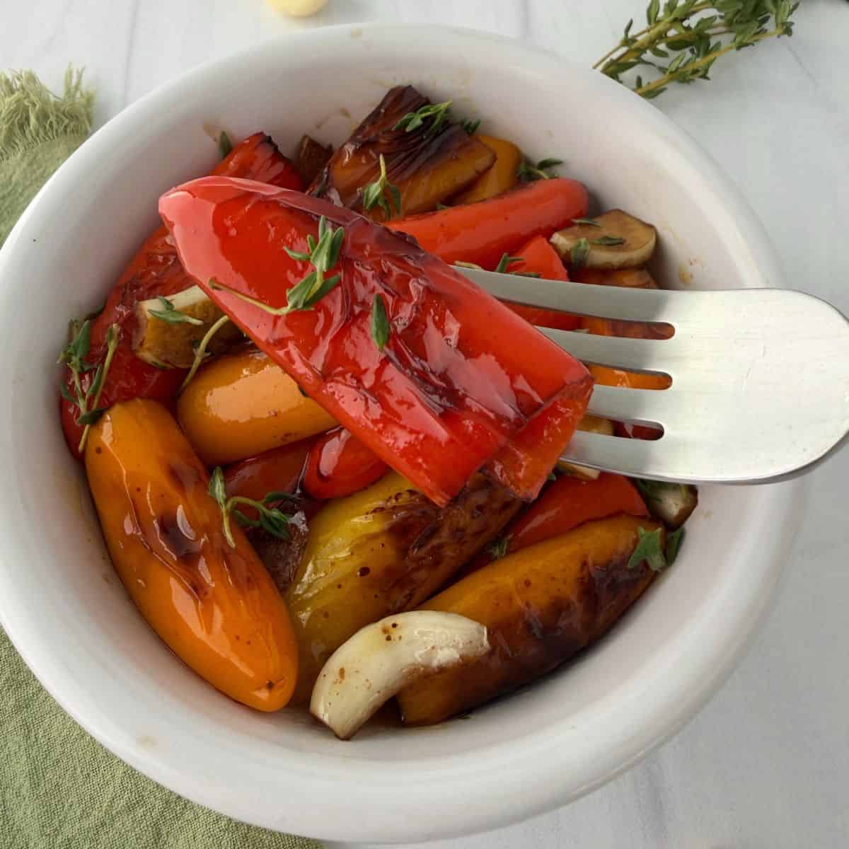 A white bowl filled with roasted mini peppers in vibrant red, yellow, and orange hues, mingled with garlic cloves and fresh herbs, as a serving fork lifts a piece of red pepper.