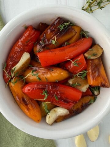 A white bowl filled with Roasted Mini Peppers-red and orange-garlic cloves, and fresh herbs, sits on a light surface beside a fork and a green napkin.