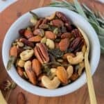 A white bowl filled with roasted nuts, including almonds, cashews, pecans, and pumpkin seeds, sits with a gold spoon beside it. Fresh herbs are placed nearby on a wooden surface.