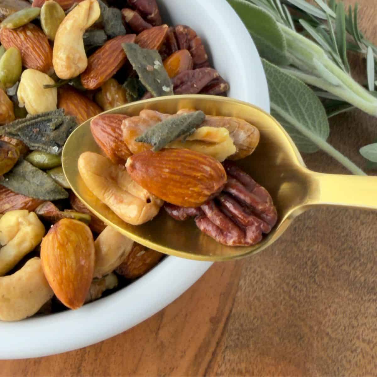 A gold spoon holds a mix of roasted nuts—almonds, pecans, cashews, pumpkin seeds—above a white bowl filled with the same blend, with fresh sage leaves in the background on a wooden surface.