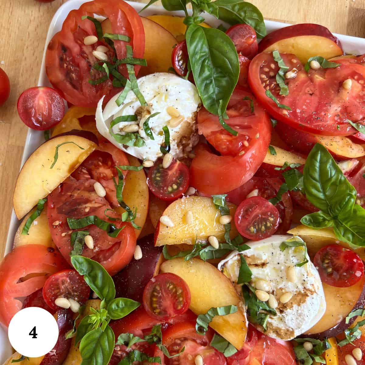 A colorful nectarine salad with sliced tomatoes, peaches, cherry tomatoes, fresh mozzarella, basil leaves, pine nuts, and a drizzle of balsamic on a white platter.