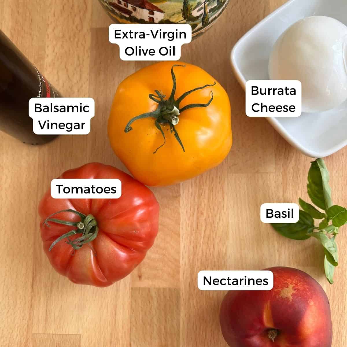 A wooden surface displays ingredients for a Nectarine Salad: a yellow tomato, red tomato, nectarine, sprig of basil, burrata in a dish, plus bottles of balsamic vinegar and extra-virgin olive oil.