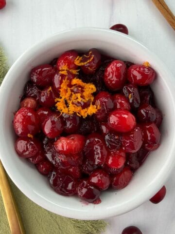 A white bowl filled with whole berry cranberry sauce, garnished with orange zest. A gold spoon rests on a green napkin next to the bowl, with cranberries and a cinnamon stick nearby on a white surface.