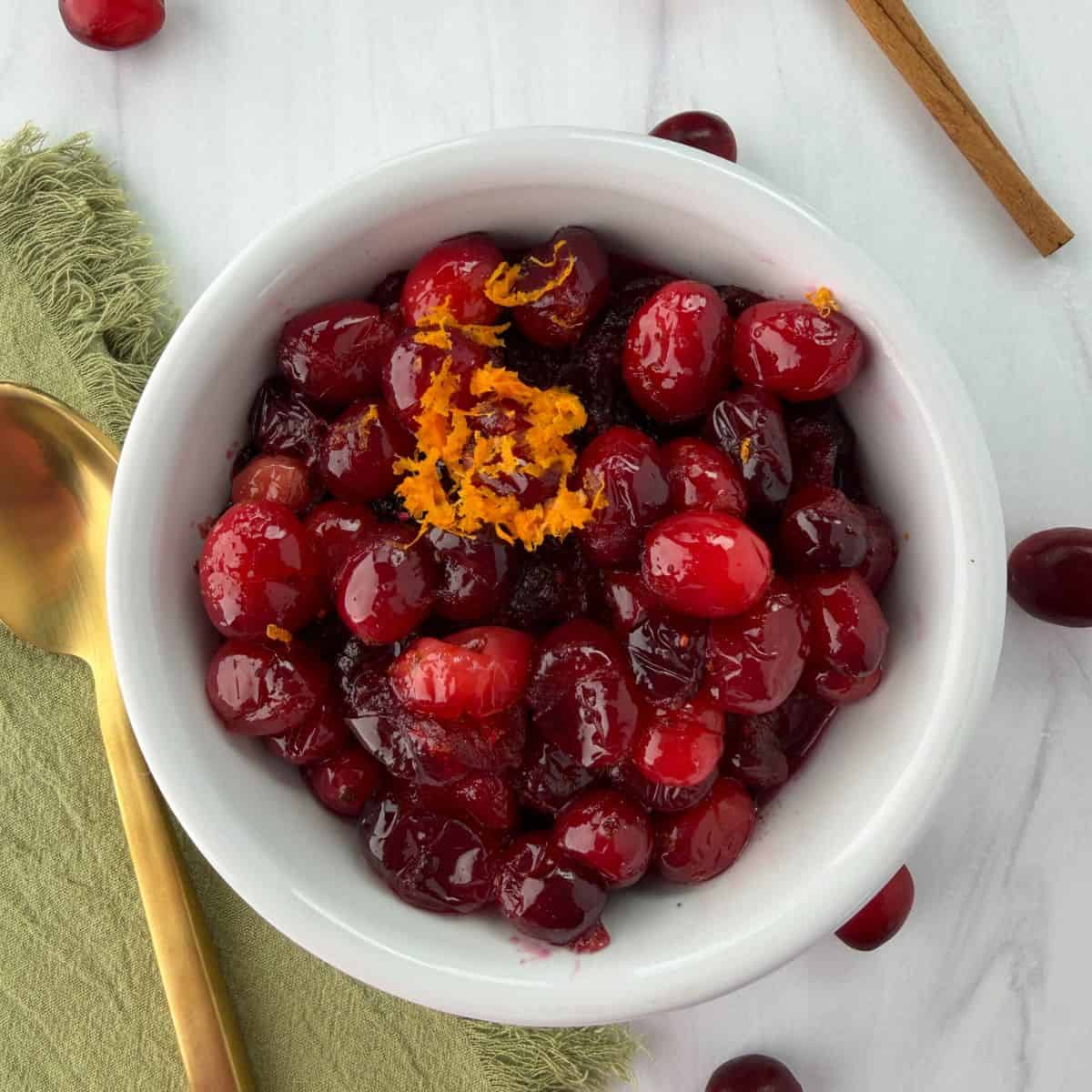 A white bowl filled with whole berry cranberry sauce, garnished with orange zest. A gold spoon rests on a green napkin next to the bowl, with cranberries and a cinnamon stick nearby on a white surface.