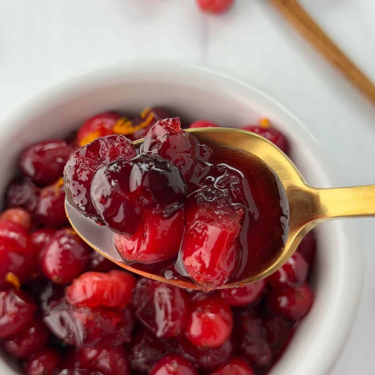 A close-up of a gold spoon holding glossy, cooked cranberries in thick whole berry cranberry sauce above a white bowl filled with more cranberries. The background is softly blurred.