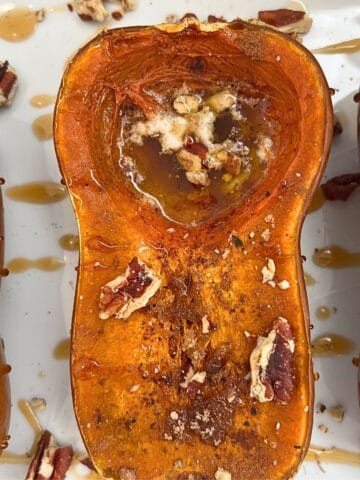 A baked roasted honeynut squash half on a white tray, topped with melted butter, chopped pecans, and a drizzle of syrup. The flesh is golden and slightly caramelized.
