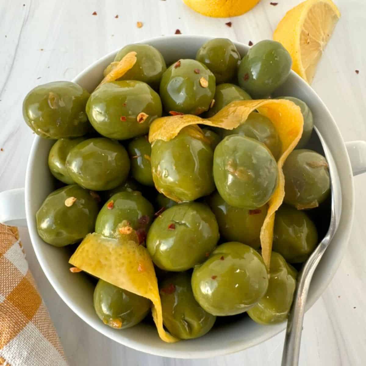 marinated olives in a white bowl with lemon peel on top. 