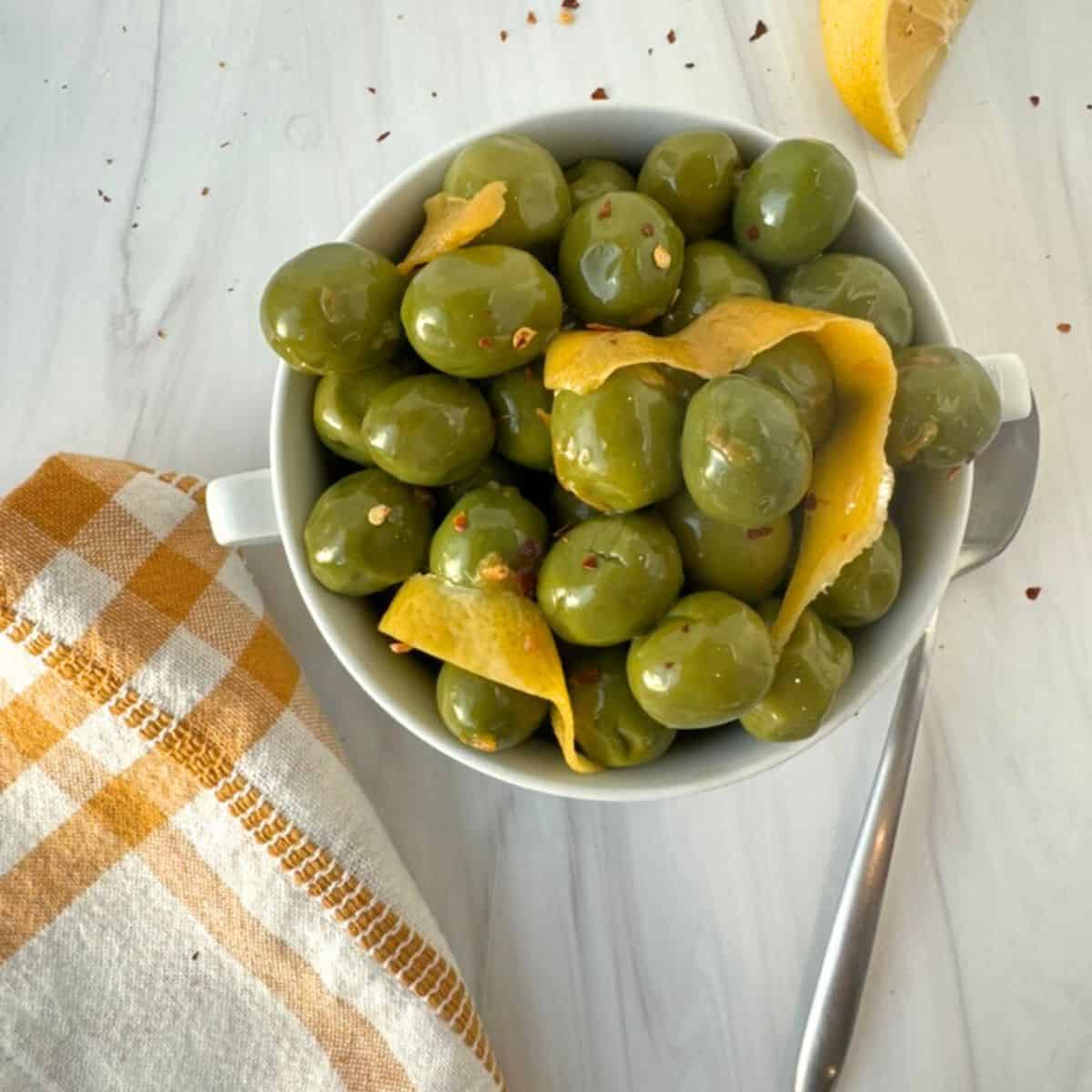 marinated olives in a white bowl with lemon peel on top and a dish towel and spoon. 