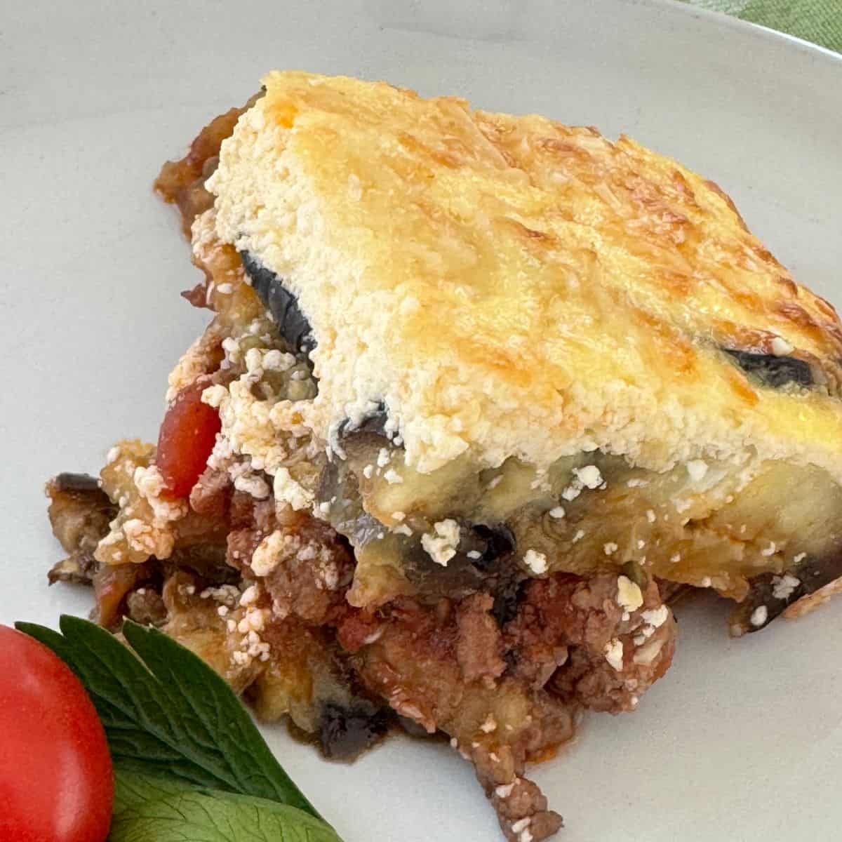 One serving of lamb moussaka on a white plate with tomatoes and parsley. 