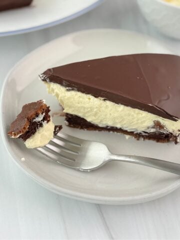 A slice of Krembo Cake with a chocolate top, creamy middle, and brownie-like base sits on a plate. A fork rests on the plate, holding a bite-sized piece. The background is a white surface.