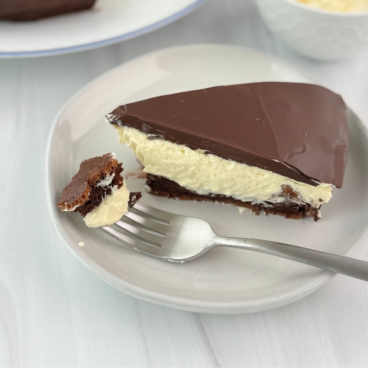 A slice of Krembo Cake with a chocolate top, creamy middle, and brownie-like base sits on a plate. A fork rests on the plate, holding a bite-sized piece. The background is a white surface.