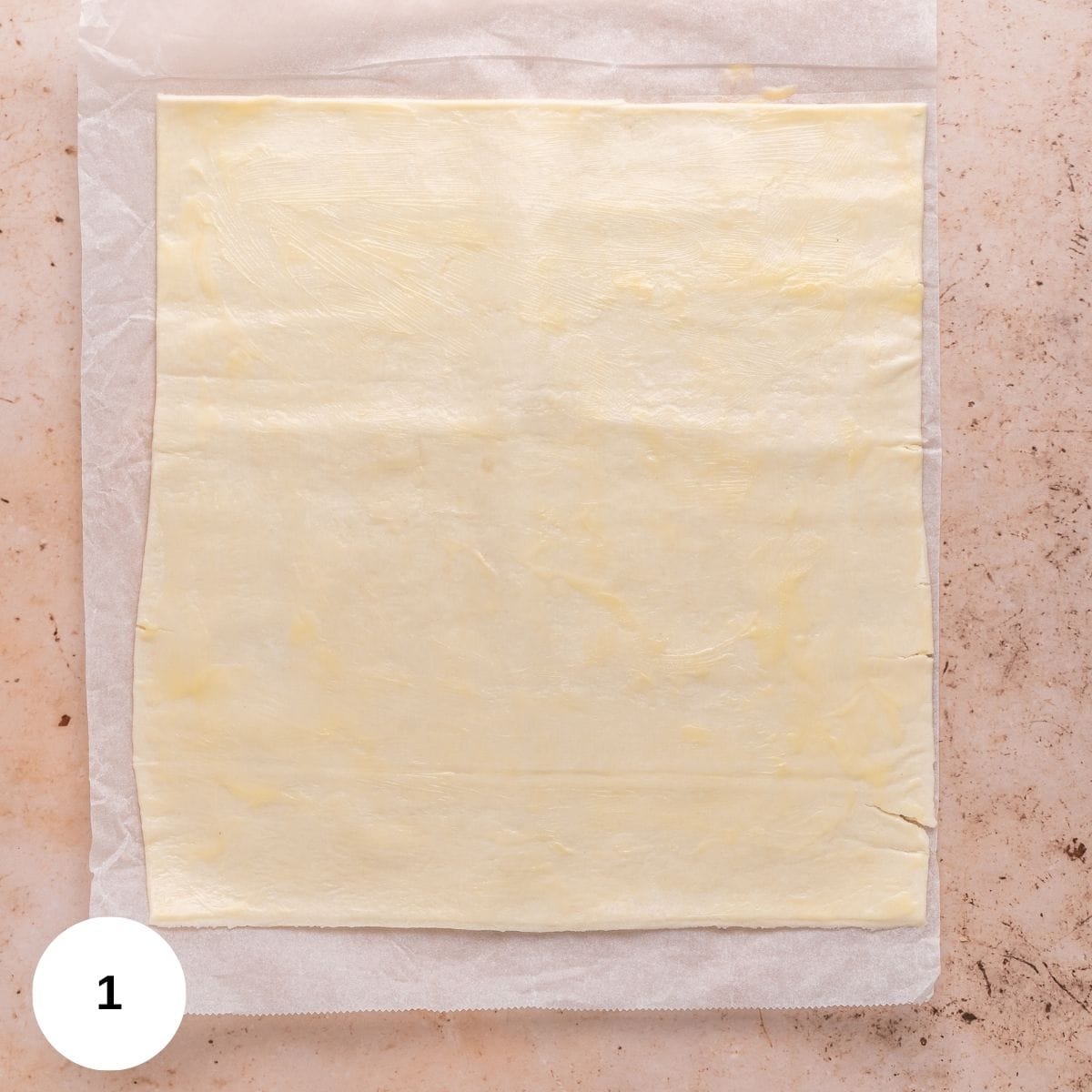 A sheet of puff pastry laid out on the counter while still sitting on parchment paper.
