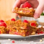 Someone picking up one raspberry white chocolate blondie from a stack of blondies on parchment paper.
