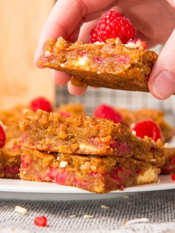 Someone picking up one raspberry white chocolate blondie from a stack of blondies on parchment paper.