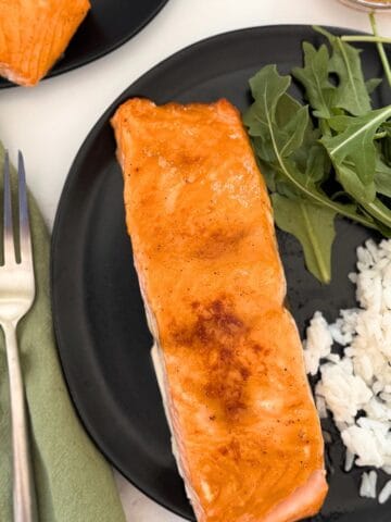 A piece of brown sugar glazed salmon on a black plate with rice and arugula.
