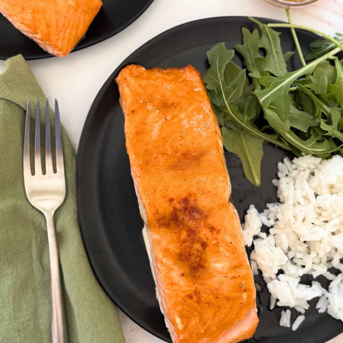 A piece of brown sugar glazed salmon on a black plate with rice and arugula.