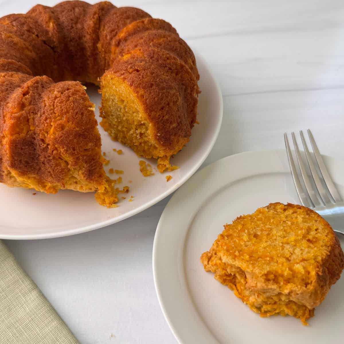 A round carrot ring bundt cake with a golden-brown crust sits on a white plate, with one slice removed. The slice rests on a separate plate beside a fork, showcasing the cake’s moist, orange interior and hint of brown sugar.