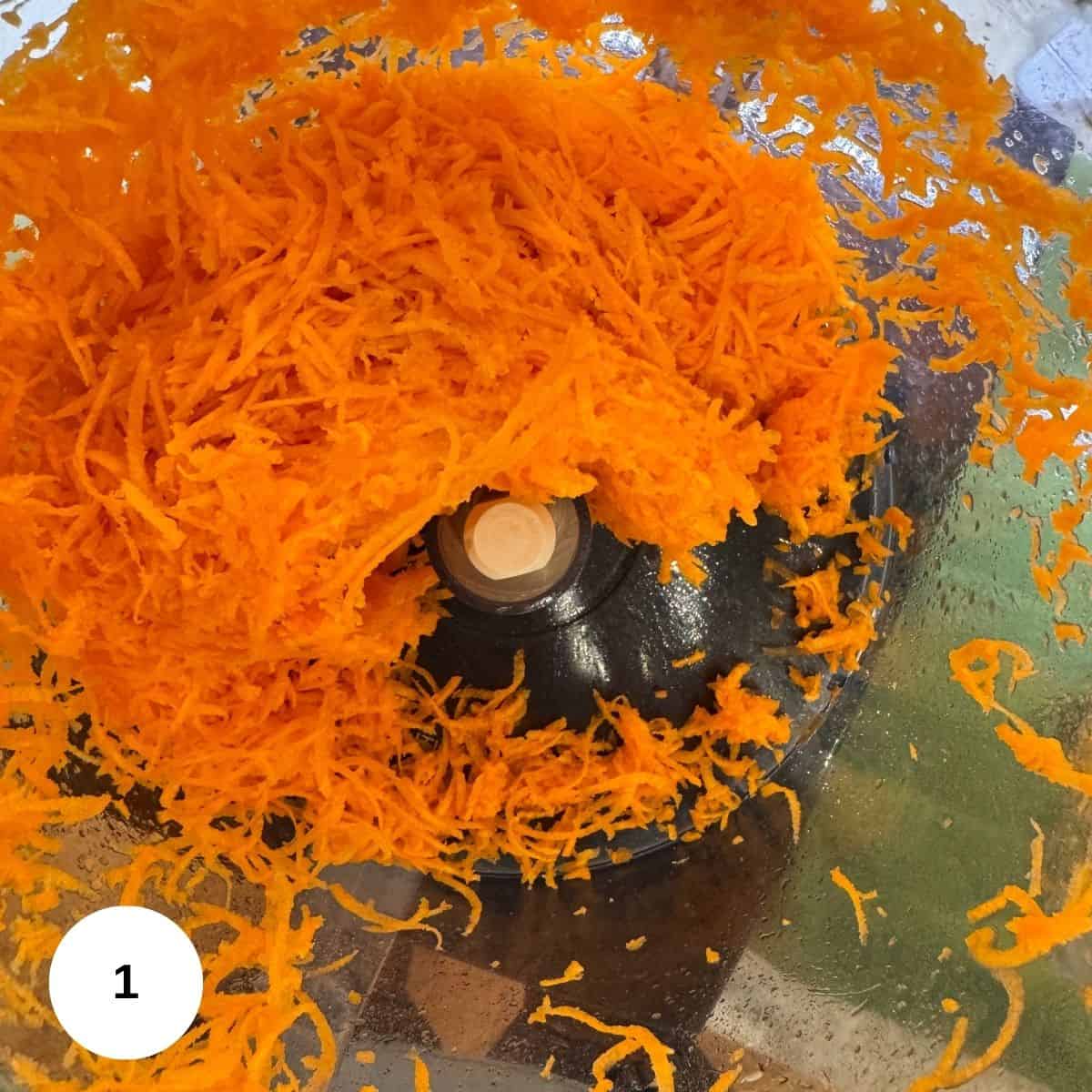 Shredded carrots collected inside a transparent food processor bowl, with some pieces sticking to the sides and the metal blade visible in the center-perfect for making a zesty carrot ring or adding a hint of lemon flavor.
