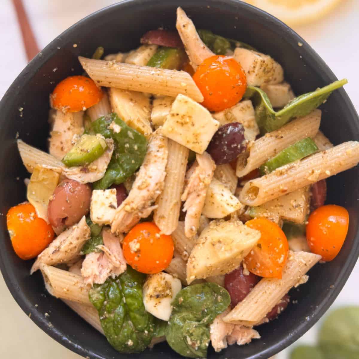 A bowl of chicken and pesto pasta salad with penne, cherry tomatoes, spinach, olives, mozzarella cubes, and chunks of chicken, all mixed with herbs and dressing.