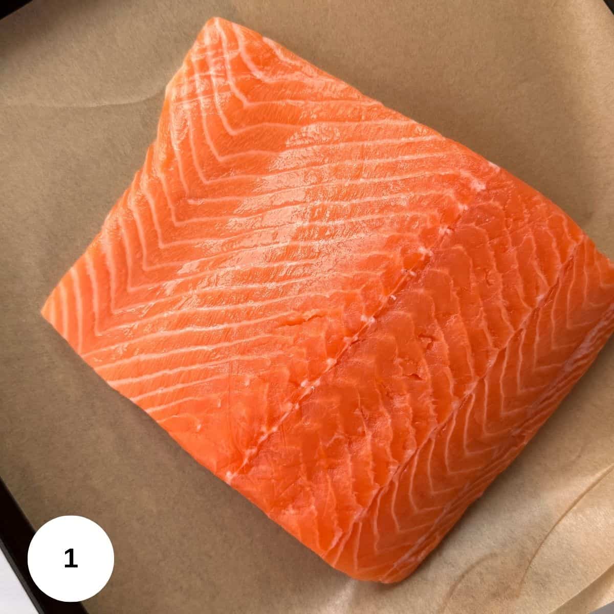 A raw, skinless fillet of salmon with visible white lines in the bright orange flesh, resting on a sheet of brown parchment paper-perfect for making fresh salmon pesto. A white circle with the number 1 appears in the lower left corner.