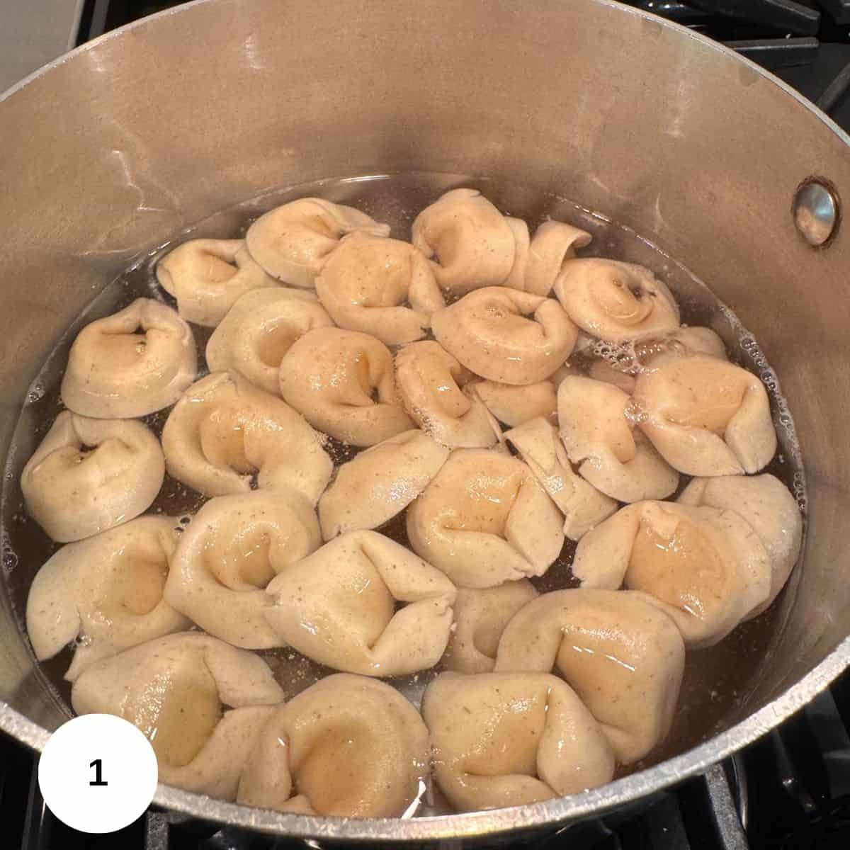 A pot of Tortellini Pesto Pasta Salad is boiling on the stove, with light brown tortellini pieces floating on the surface of the bubbling water.