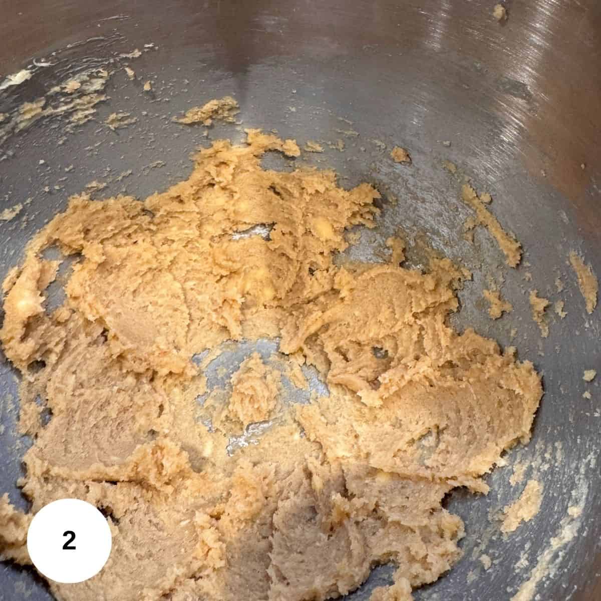 Creamed butter and brown sugar mixture in a metal mixing bowl, with some of the bowl sides visible. A white circle with the number 2 is in the bottom left corner.