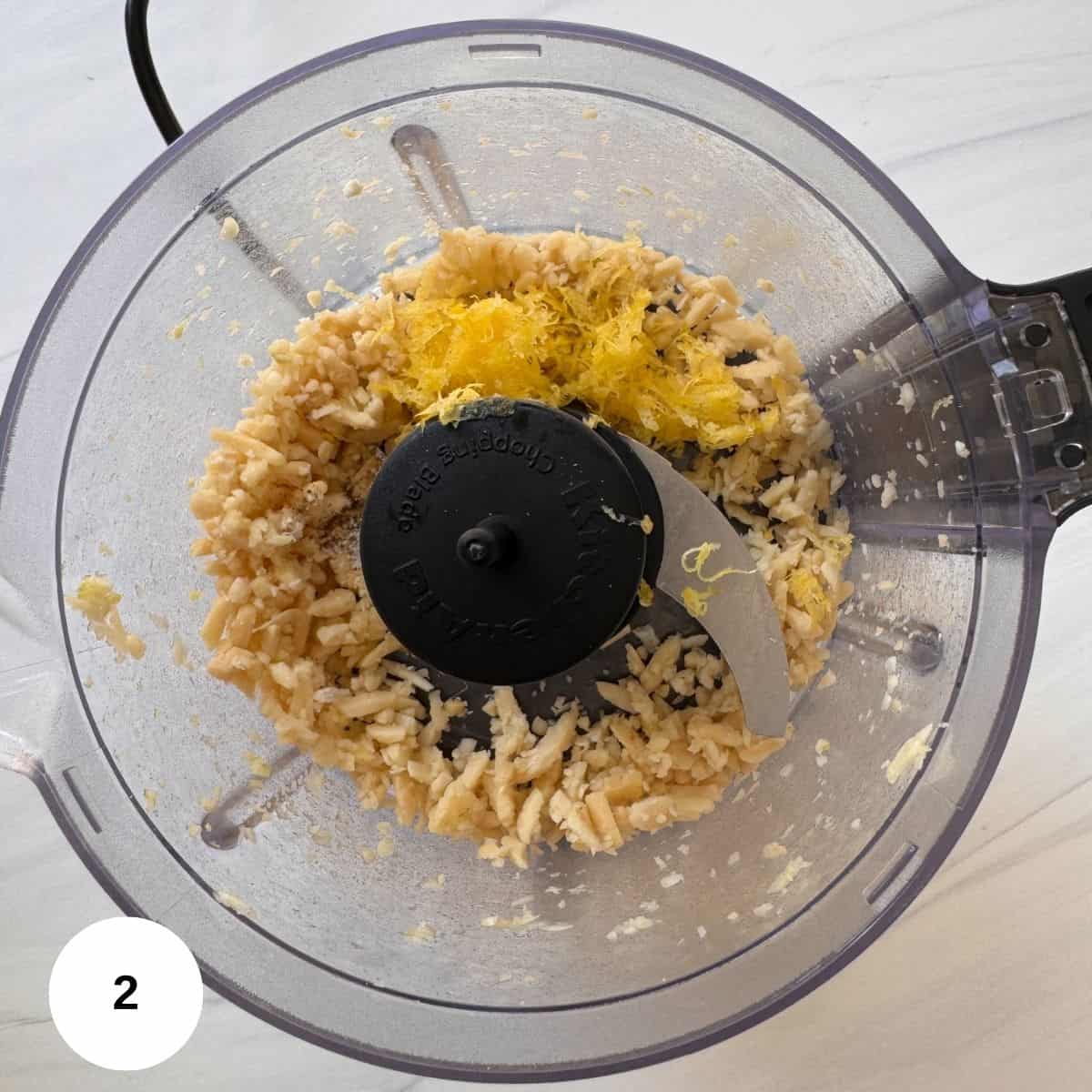 A food processor bowl containing blended cashews and a pile of grated lemon zest, ideal for making mint pesto, viewed from above. The blade is visible, and a white circle with the number 2 is in the bottom left corner.