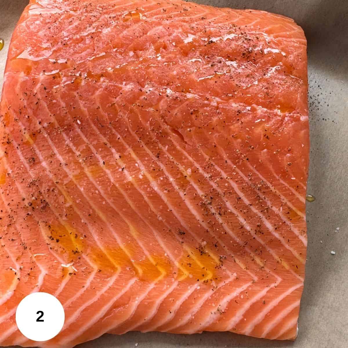 A close-up of a raw salmon fillet on parchment paper, seasoned with salt, pepper, and drizzled with oil-perfect for making salmon pesto. The bright orange flesh and marbling are visible. A small white circle with the number 2 is in the bottom left corner.