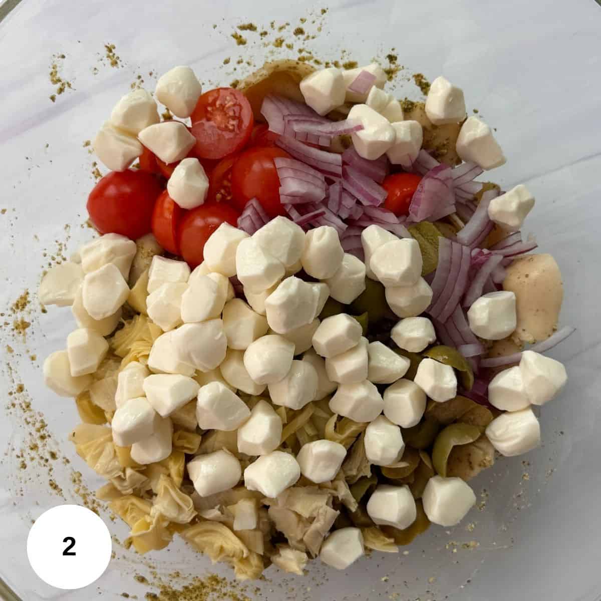A glass bowl filled with cherry tomatoes, sliced red onions, artichoke hearts, green olives, and small mozzarella balls-perfect ingredients for a fresh Tortellini Pesto Pasta Salad-layered and ready to mix. A white circle with the number 2 is in the bottom left corner.