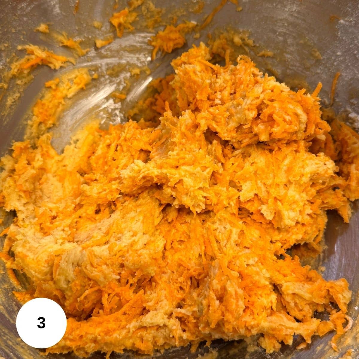 A close-up of orange and beige shredded mixture in a mixing bowl, likely a batter for Carrot Ring containing grated carrots and brown sugar. A white label with the number 3 appears in the lower left corner.