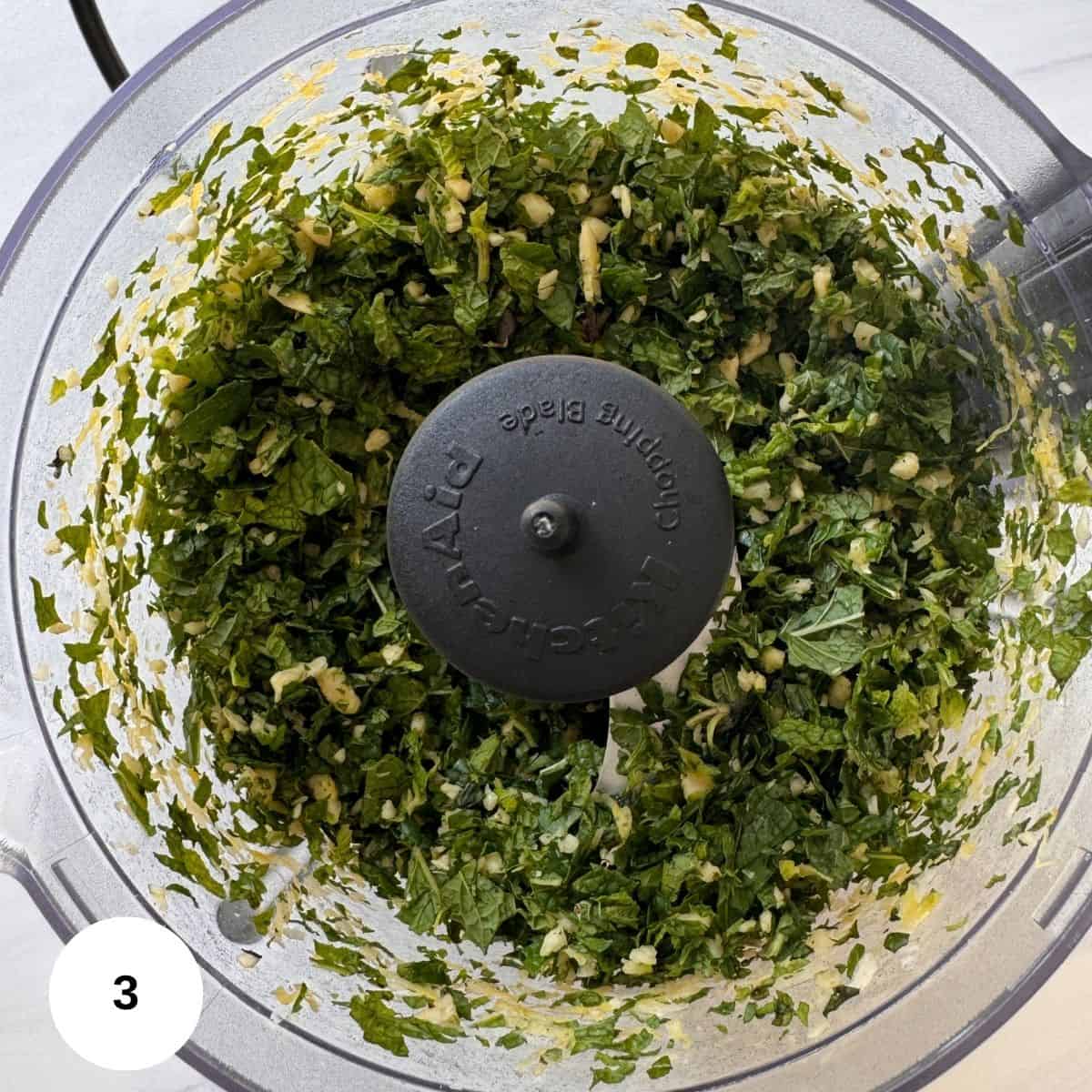 Chopped green herbs and garlic combine for a fresh mint pesto in a food processor bowl, viewed from above. A black blade attachment is visible in the center. A white circle with the number 3 appears in the lower left corner.