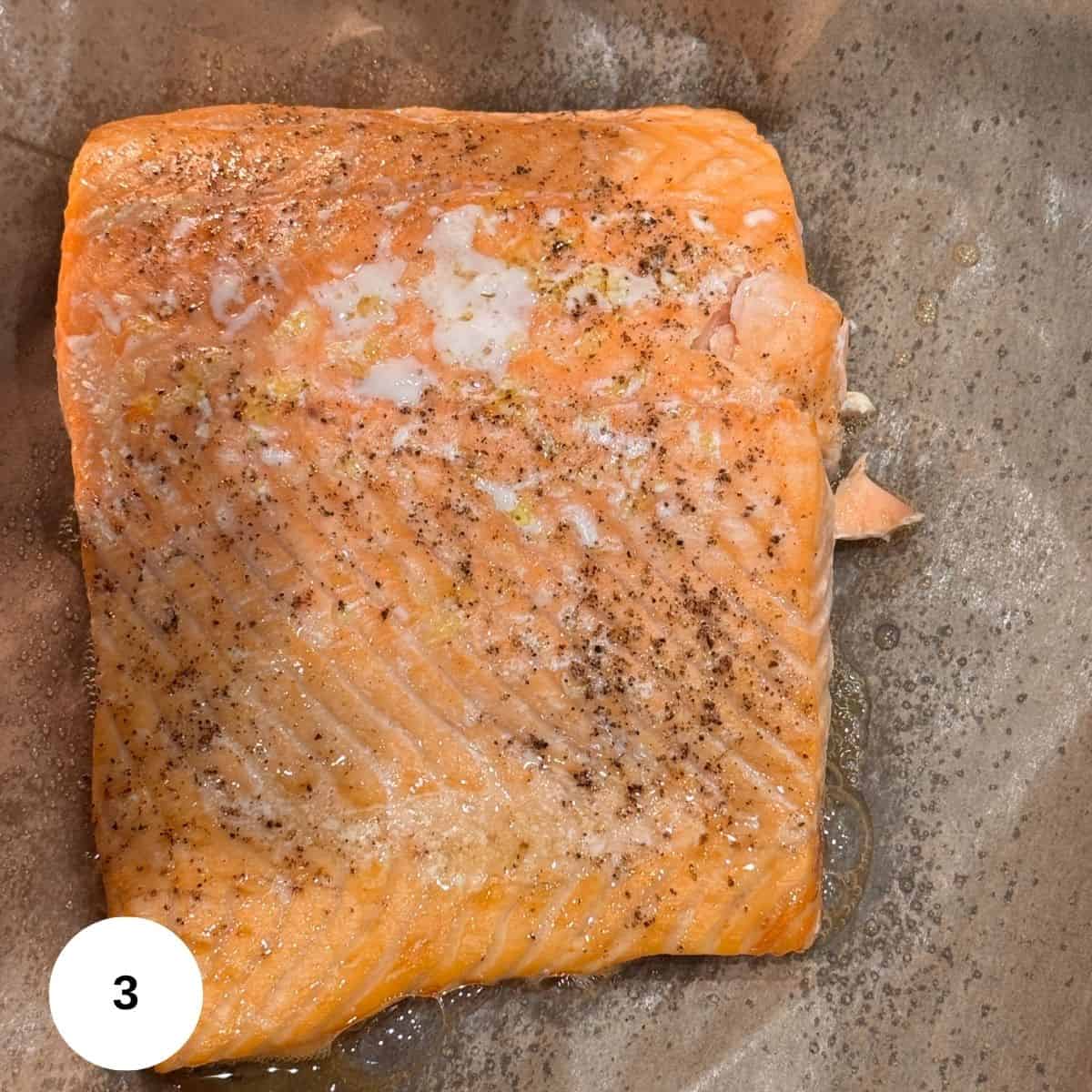 A cooked salmon pesto fillet seasoned with black pepper and salt rests on brown parchment paper, with a small amount of oil around it. A white circle with the number 3 appears in the lower left corner.