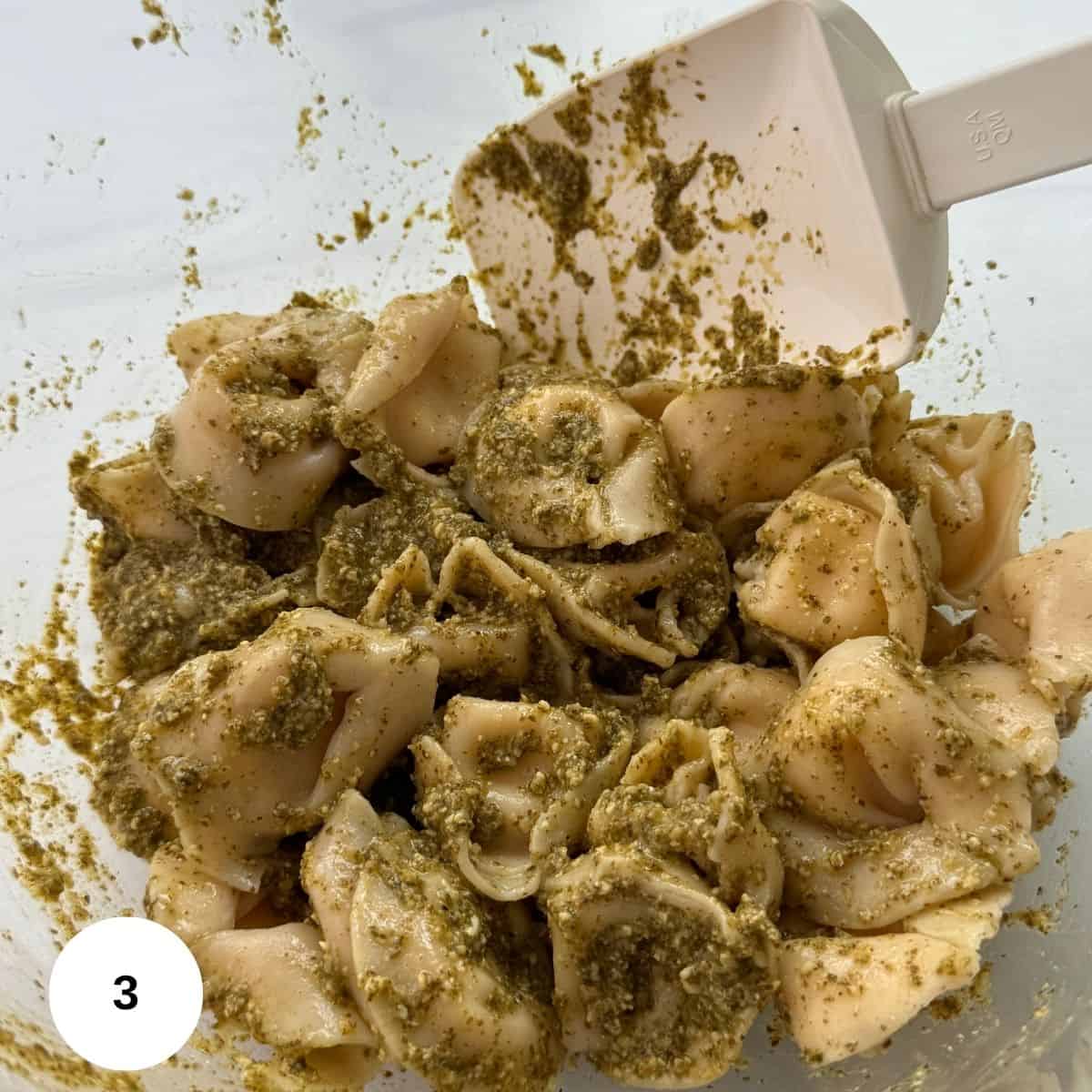 A close-up of Tortellini Pesto Pasta Salad, with tortellini coated in green pesto sauce, being mixed with a white spatula inside a clear bowl. A white circle with the number 3 is in the lower left corner.