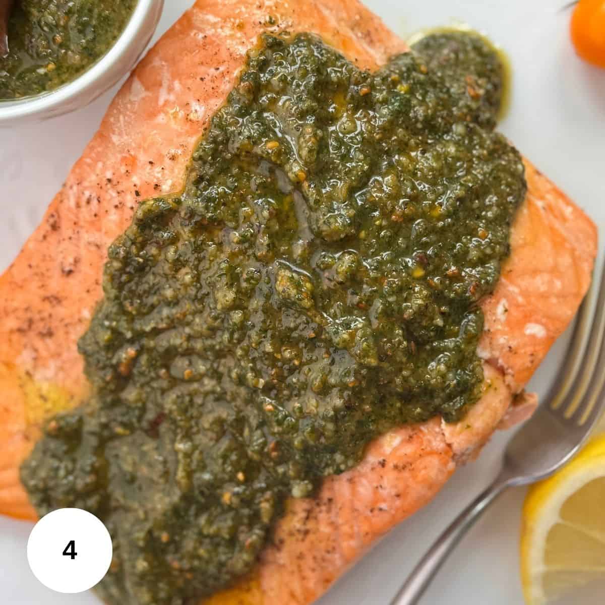A cooked salmon fillet topped with vibrant salmon pesto sauce is plated next to a lemon wedge, cherry tomato, and a small bowl of extra sauce. A fork rests at the side.