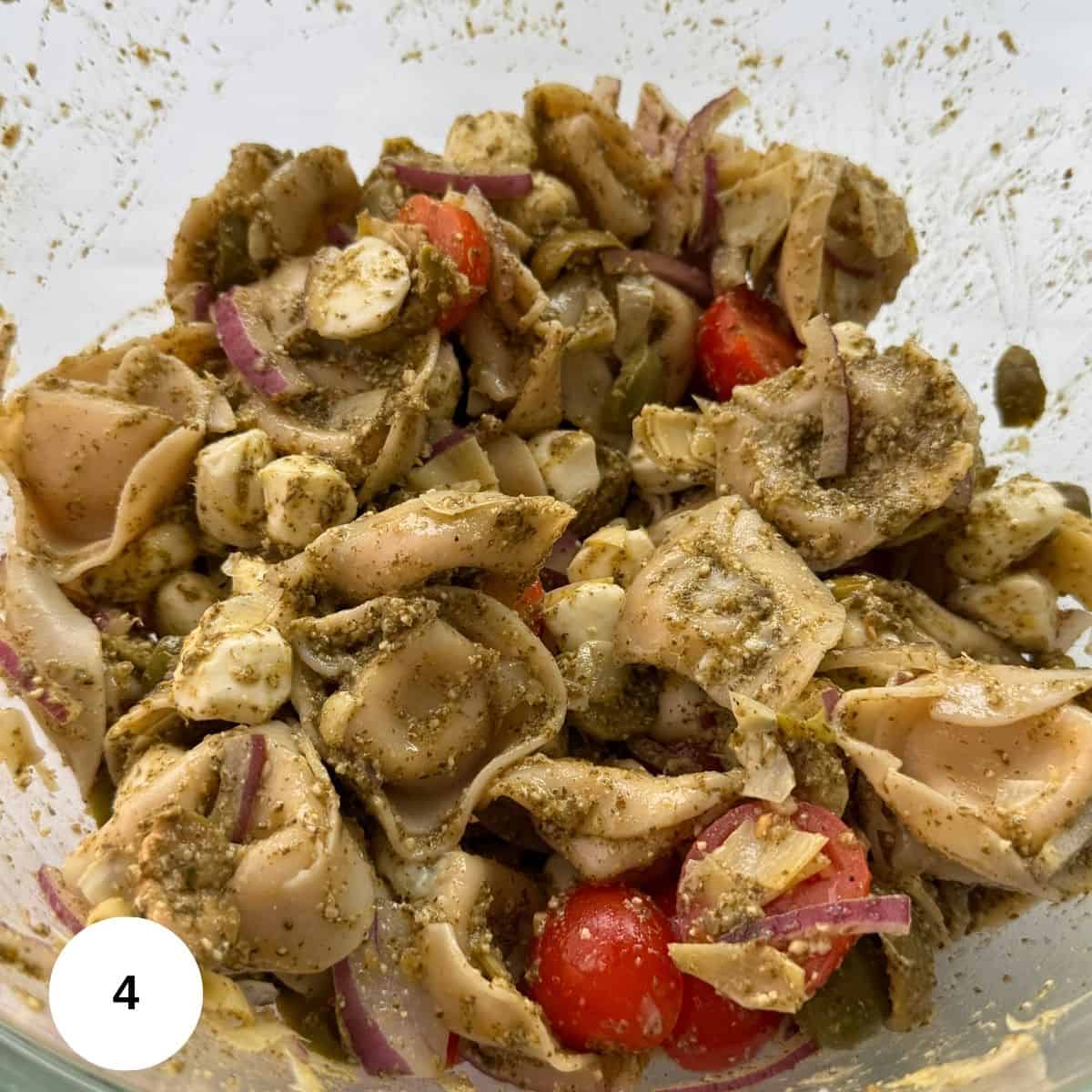 A close-up of Tortellini Pesto Pasta Salad with cherry tomatoes, red onion slices, artichoke hearts, and mozzarella balls in a clear bowl. A white circle with the number 4 is in the lower left corner.