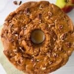 A round Apple Caramel Cake topped with chopped pecans sits on a light surface with an apple nearby. The cake has a golden-brown, rustic appearance, a moist center, and is perfect drizzled with caramel glaze.