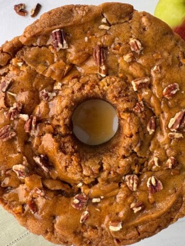 A round Apple Caramel Cake topped with chopped pecans sits on a light surface with an apple nearby. The cake has a golden-brown, rustic appearance, a moist center, and is perfect drizzled with caramel glaze.