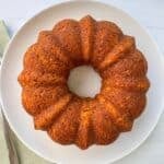 A golden-brown carrot ring Bundt cake with a ridged, ring shape sits on a white plate atop a light-colored surface.