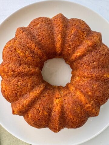 A golden-brown carrot ring Bundt cake with a ridged, ring shape sits on a white plate atop a light-colored surface.