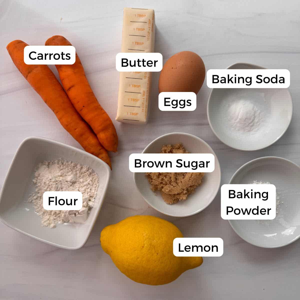 Two carrots, a stick of butter, an egg, brown sugar, flour, baking soda, baking powder, and a lemon are arranged on a white surface. Each item is labeled—perfect ingredients for a delicious carrot ring dessert.