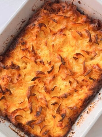 A baked cottage cheese noodle casserole with a golden, crispy, and slightly browned top layer, featuring swirls and peaks in a white rectangular dish. The edges show some caramelization and browning.