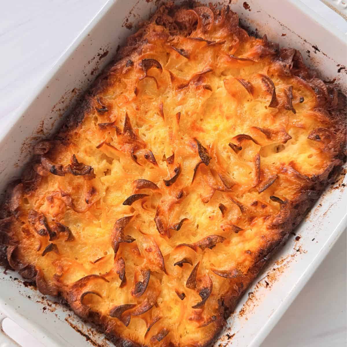 A baked cottage cheese noodle casserole with a golden, crispy, and slightly browned top layer, featuring swirls and peaks in a white rectangular dish. The edges show some caramelization and browning.