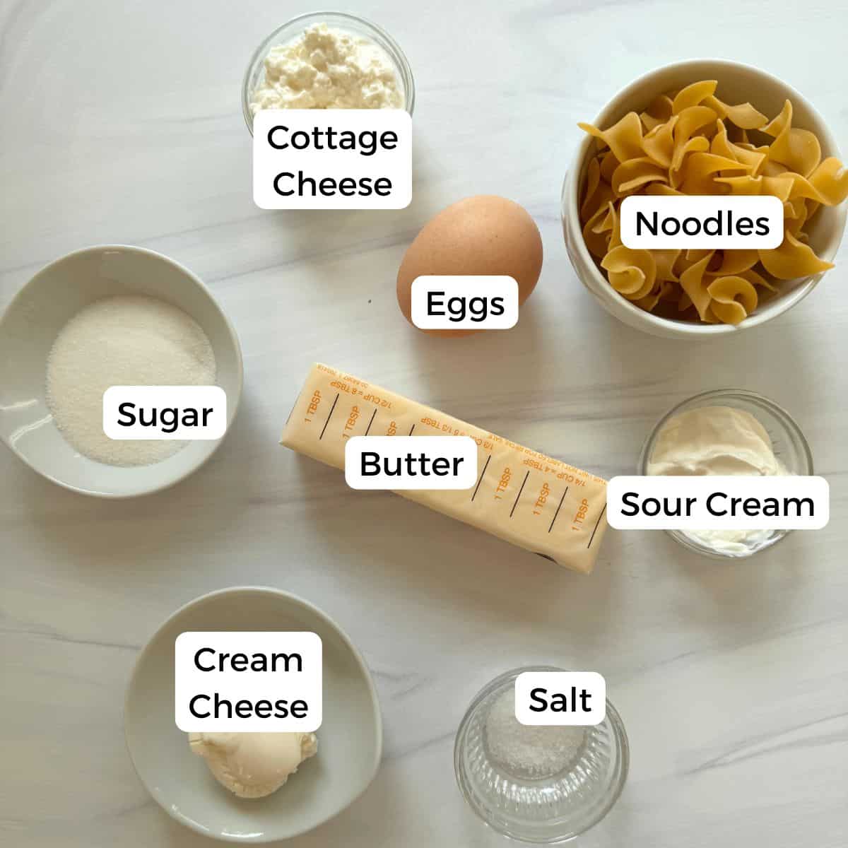 A flat lay of labeled ingredients for cottage cheese noodle: cottage cheese, an egg, noodles, sugar, butter, sour cream, cream cheese, and salt on a light surface.