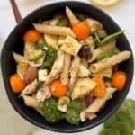 A black bowl filled with chicken and pesto pasta salad-penne tossed with cherry tomatoes, spinach leaves, mozzarella chunks, olives, and chicken in pesto sauce-sits on a light surface.