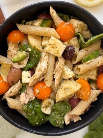 A black bowl filled with chicken and pesto pasta salad—penne tossed with cherry tomatoes, spinach leaves, mozzarella chunks, olives, and chicken in pesto sauce—sits on a light surface.
