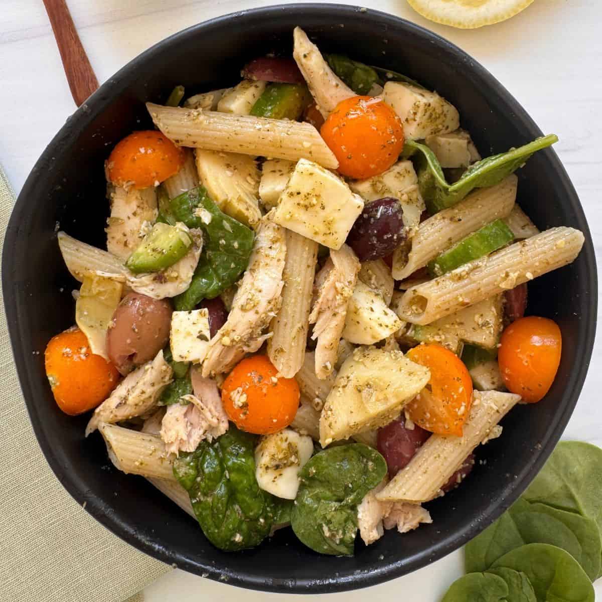 A black bowl filled with chicken and pesto pasta salad-penne tossed with cherry tomatoes, spinach leaves, mozzarella chunks, olives, and chicken in pesto sauce-sits on a light surface.