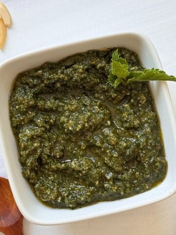 A square white bowl filled with vibrant mint pesto sauce, garnished with a sprig of mint. A wooden spoon and slivered almonds rest nearby on a white surface.