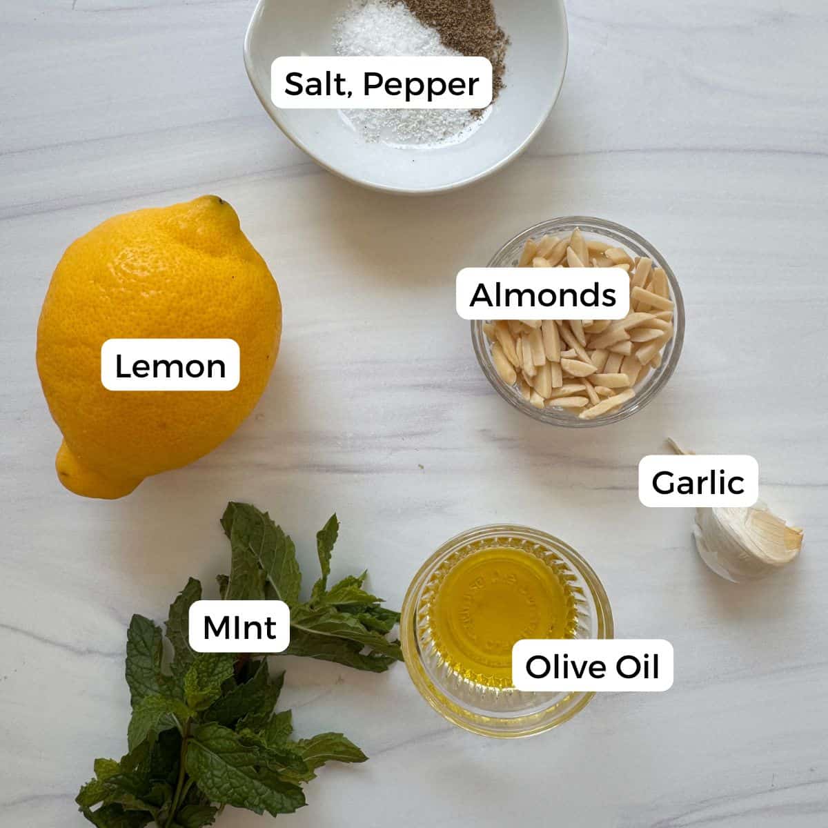 Ingredients for mint pesto on a white surface: a lemon, fresh mint leaves, a small bowl of slivered almonds, garlic, salt and pepper, and olive oil. Each item is labeled for easy preparation.