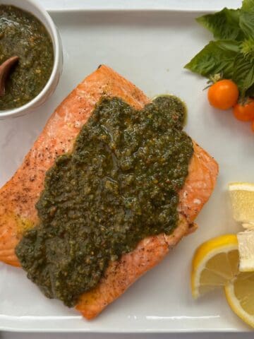 A cooked salmon pesto fillet topped with green sauce on a white plate, garnished with lemon wedges, yellow cherry tomatoes, fresh basil, and a small bowl of extra pesto with a wooden spoon.