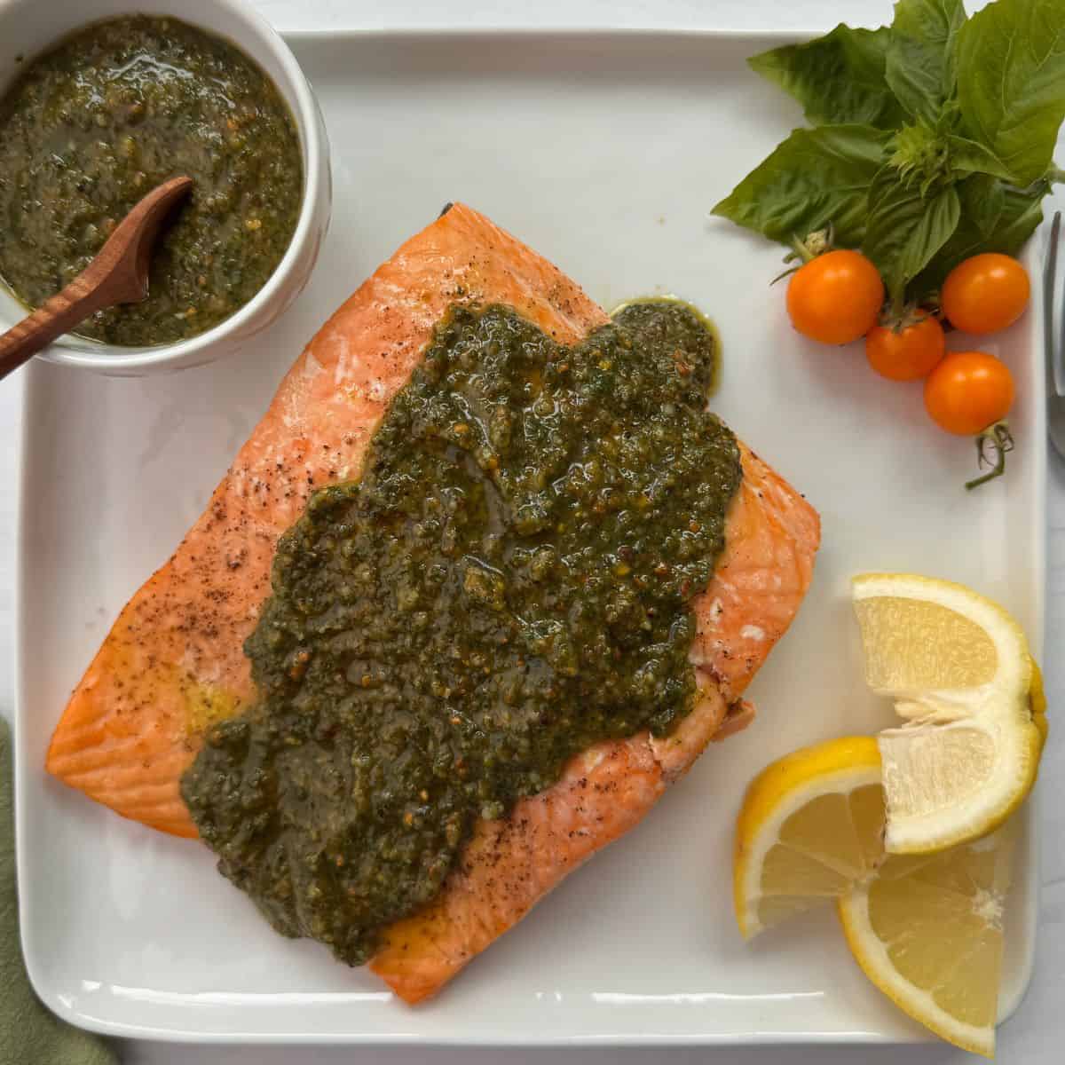 A cooked salmon pesto fillet topped with green sauce on a white plate, garnished with lemon wedges, yellow cherry tomatoes, fresh basil, and a small bowl of extra pesto with a wooden spoon.